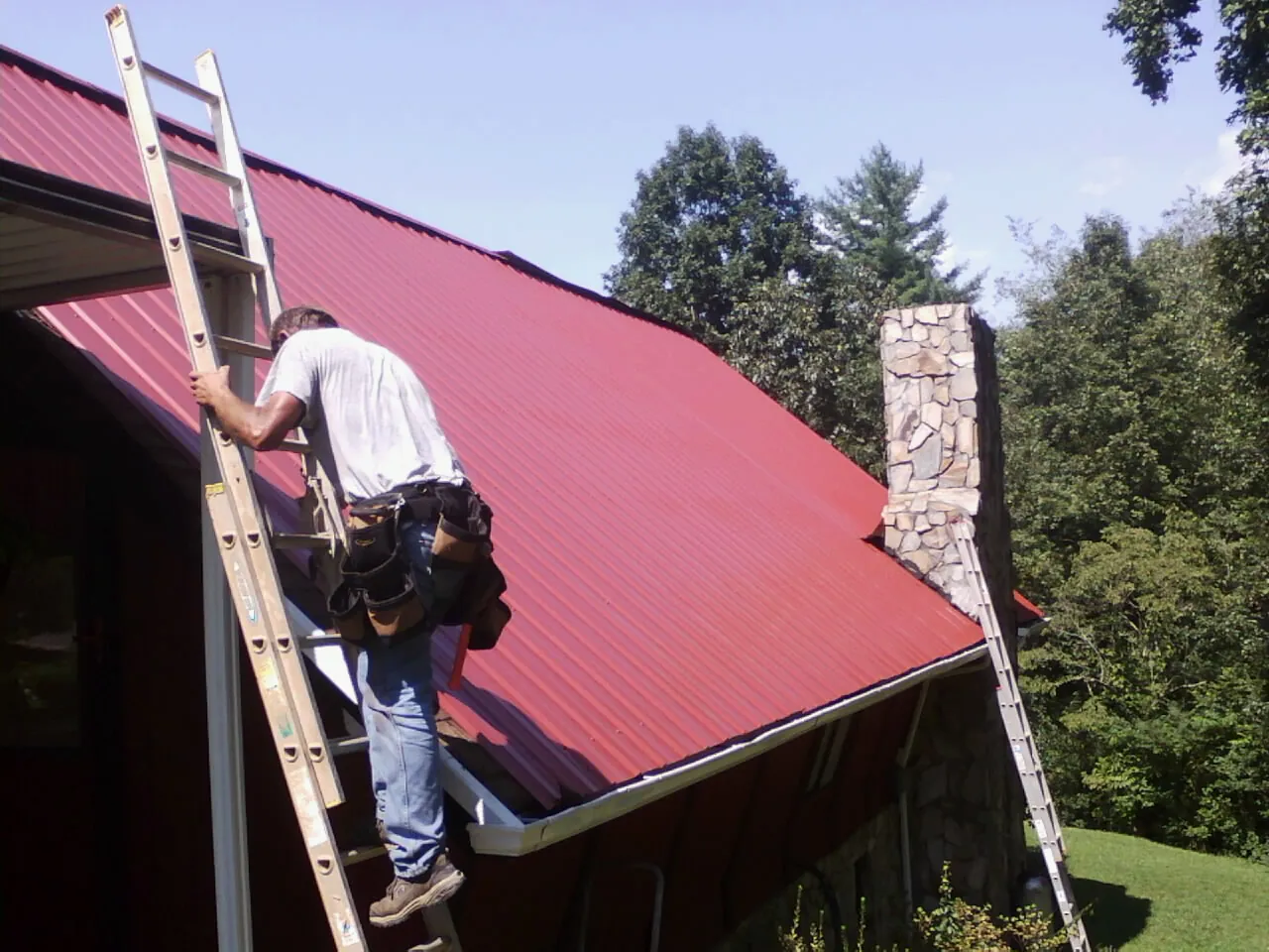 Roofer on a ladder beside a red metal roof and stone chimney — Soffit & Fascia Repair by Roofing Brothers in Topton